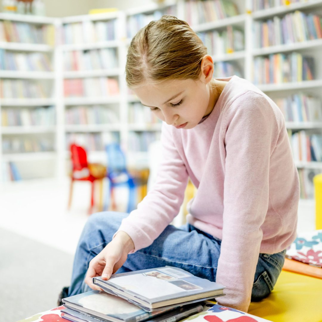 Pretty girl child reading book in library. Cute female preteen kid studying literature and making research for school in biblioteque